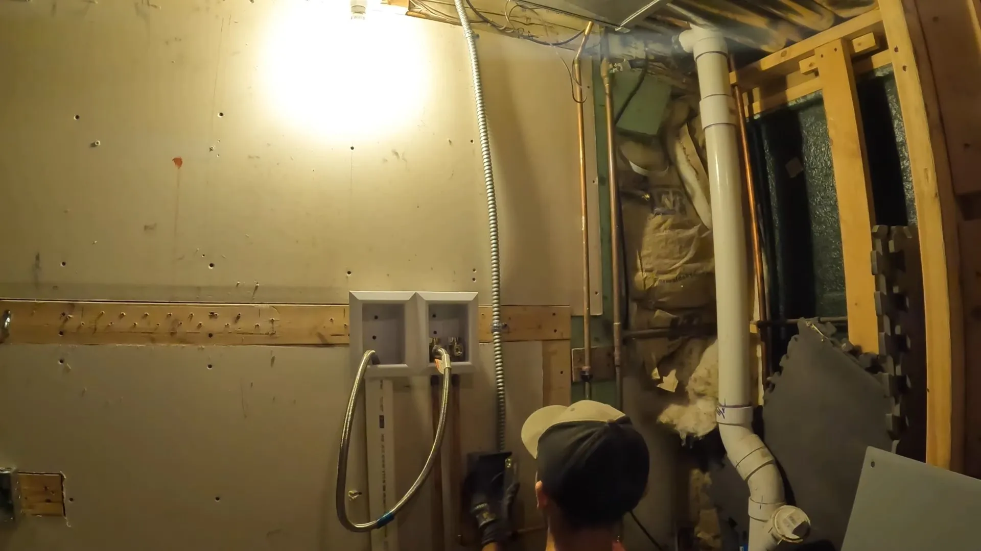 Basement wall opened to the framing showing a laundry supply box, a white drain pipe and wiring