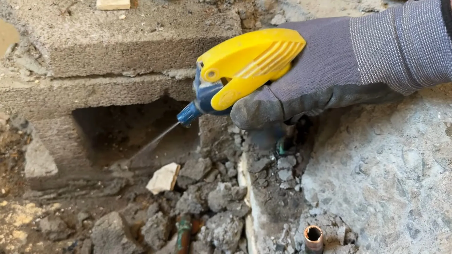 Gloved hand holding a yellow spray nozzle over a broken concrete slab with a copper stub showing