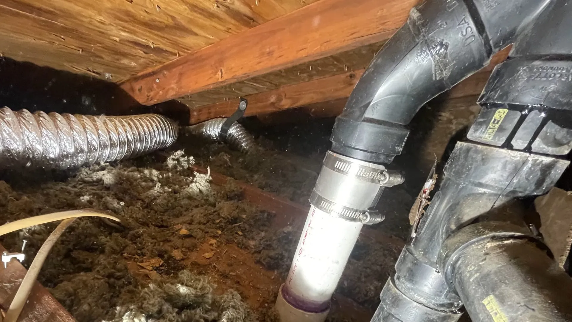 Gray drain fittings and a flexible duct under floor joists in a crawl space with debris below