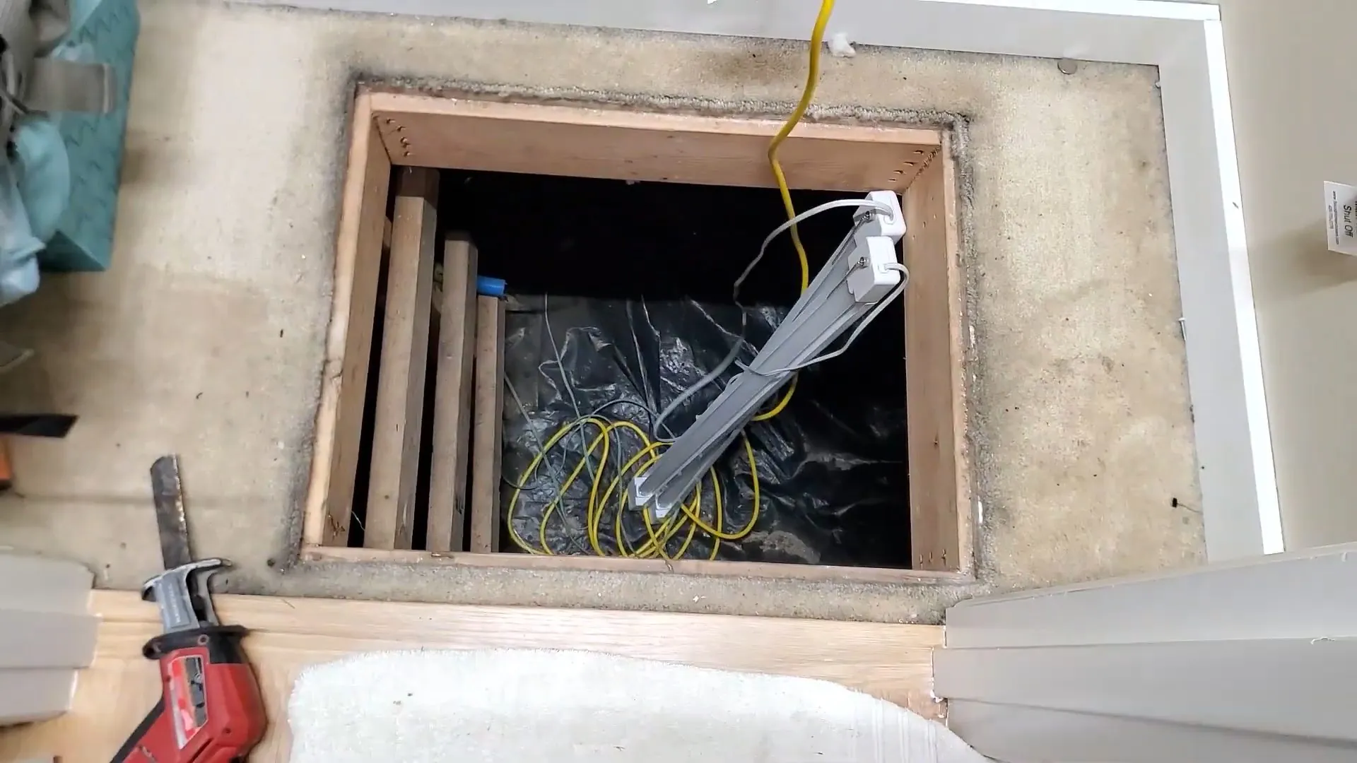 Square access opening cut into a floor above a crawl space lined with black plastic and a work light