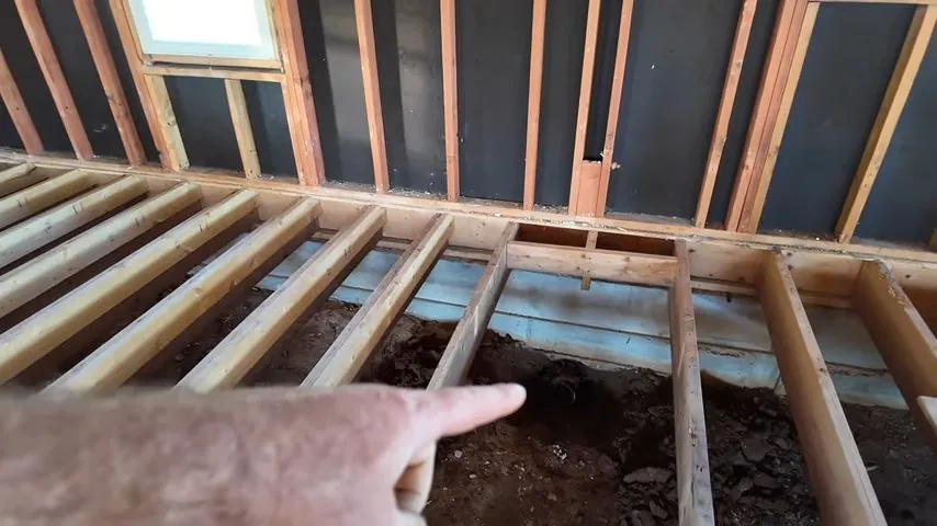 Floor joists exposed above bare soil with a hand pointing at the ground below