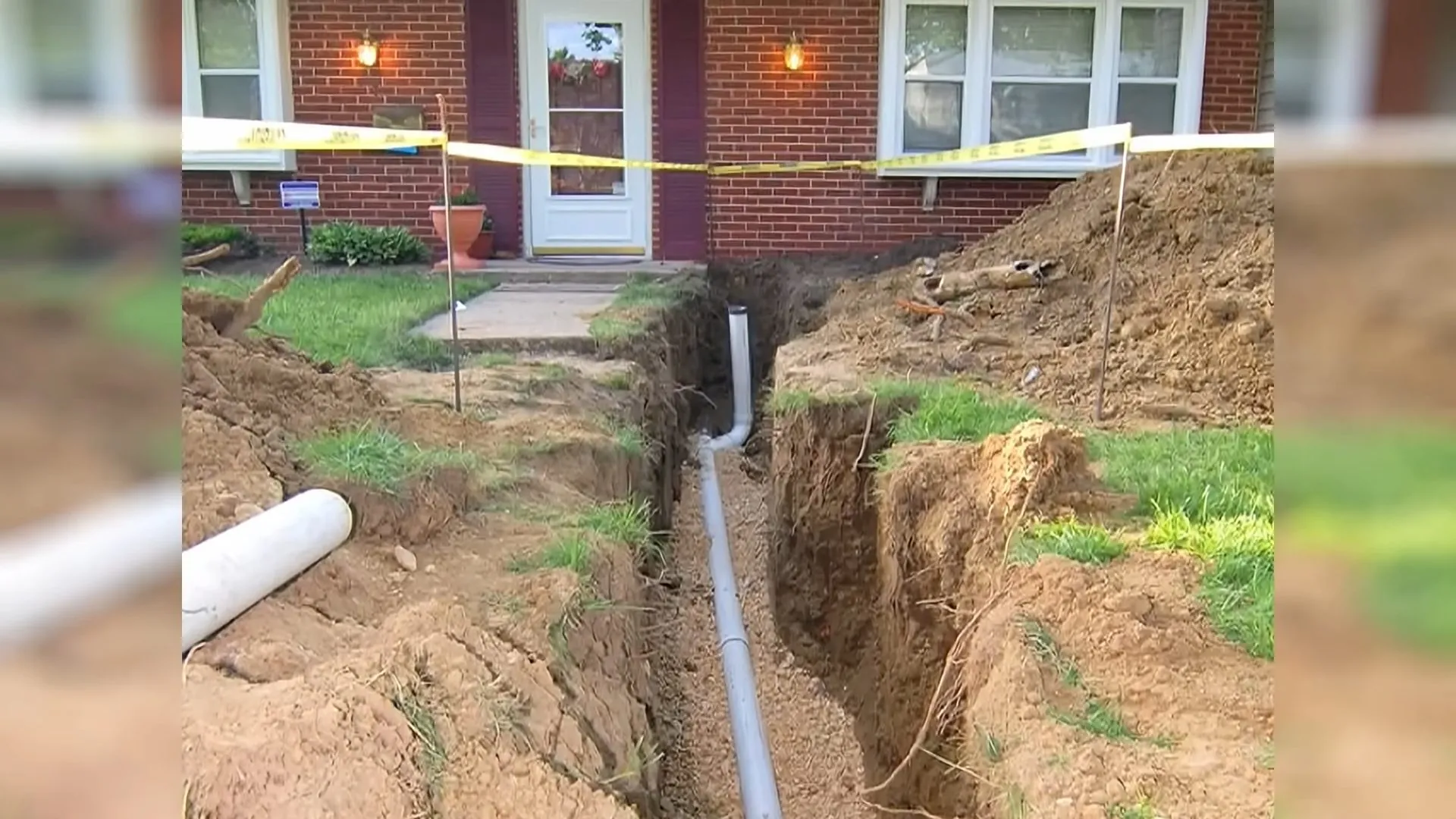 Trench across a front lawn with gray pipe laid in it and caution tape in front of a brick house