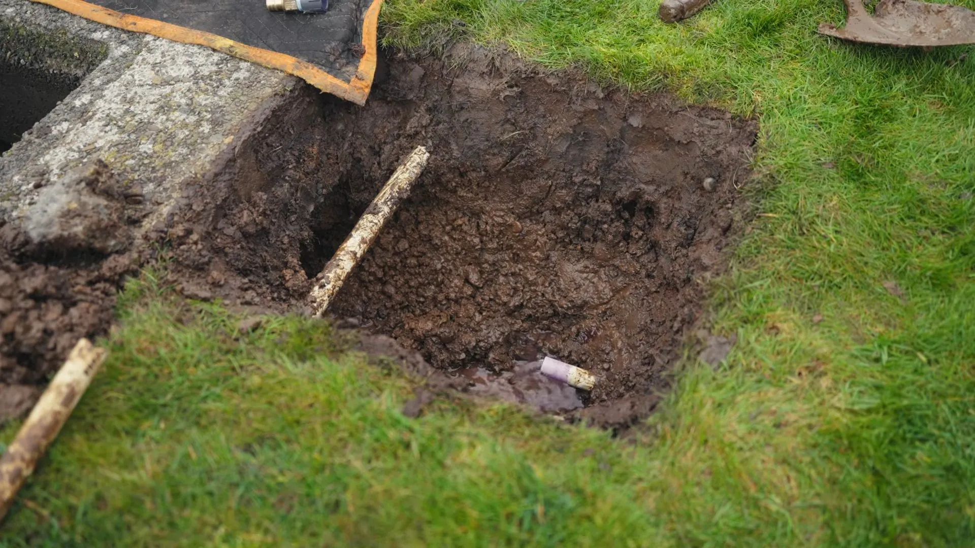 Shovel beside a hand-dug hole in a lawn exposing a buried supply line