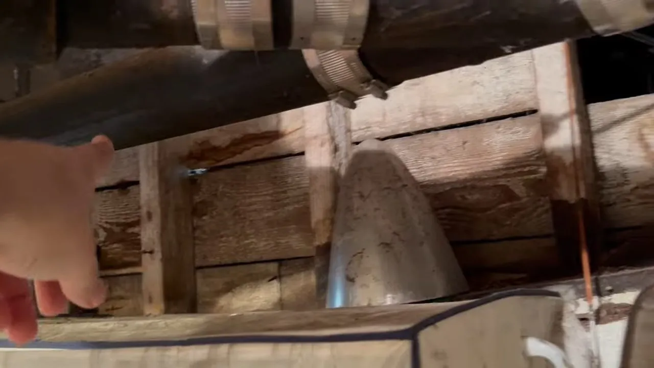 Old black cast iron drain pipe running under wood subfloor boards with a hand reaching in