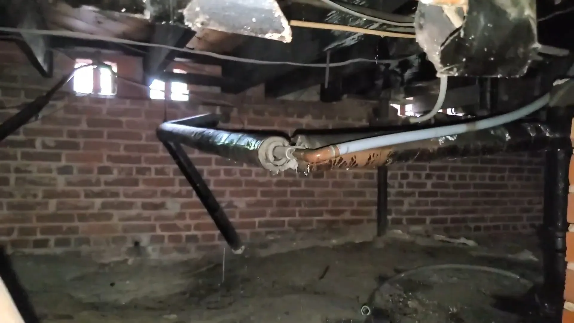 Old drain and supply piping under a floor in a brick-walled crawlspace