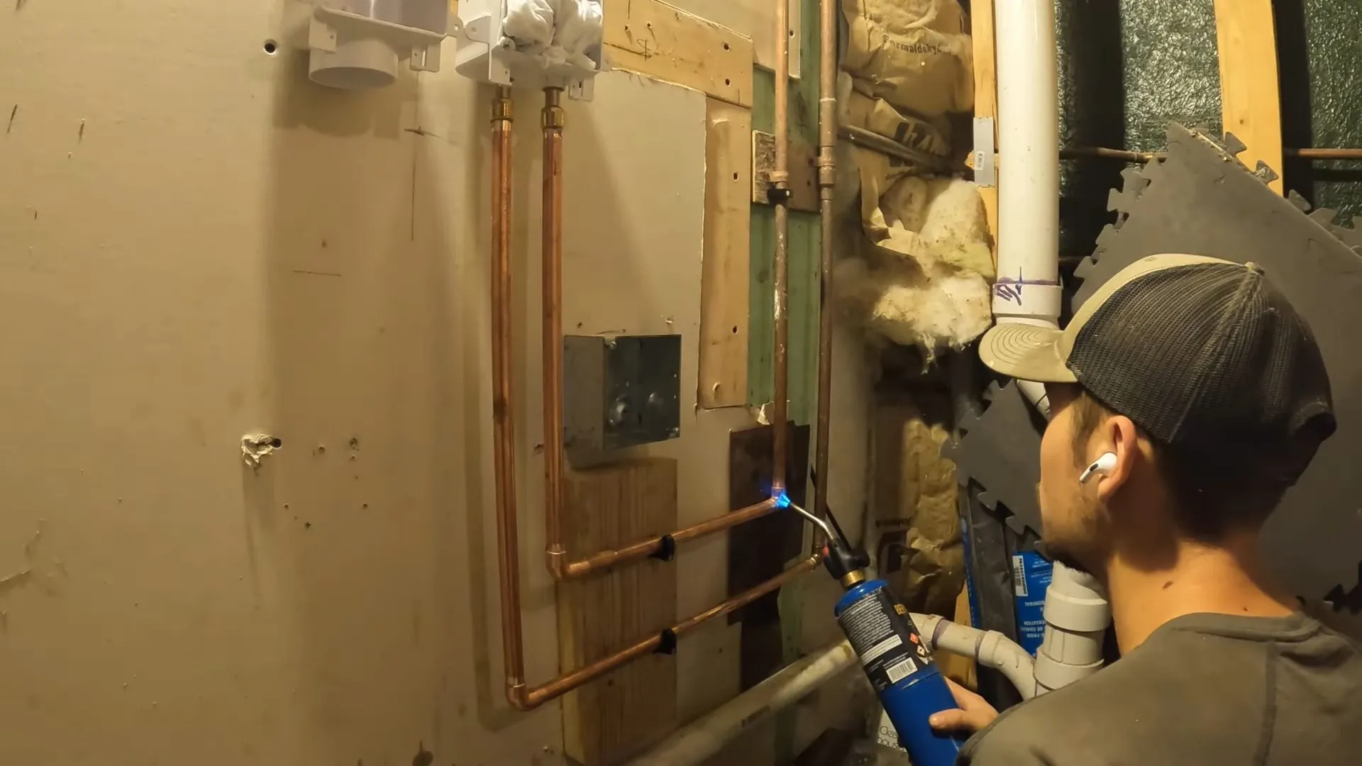 Plumber with a torch soldering copper lines on a basement wall beside a white drain pipe