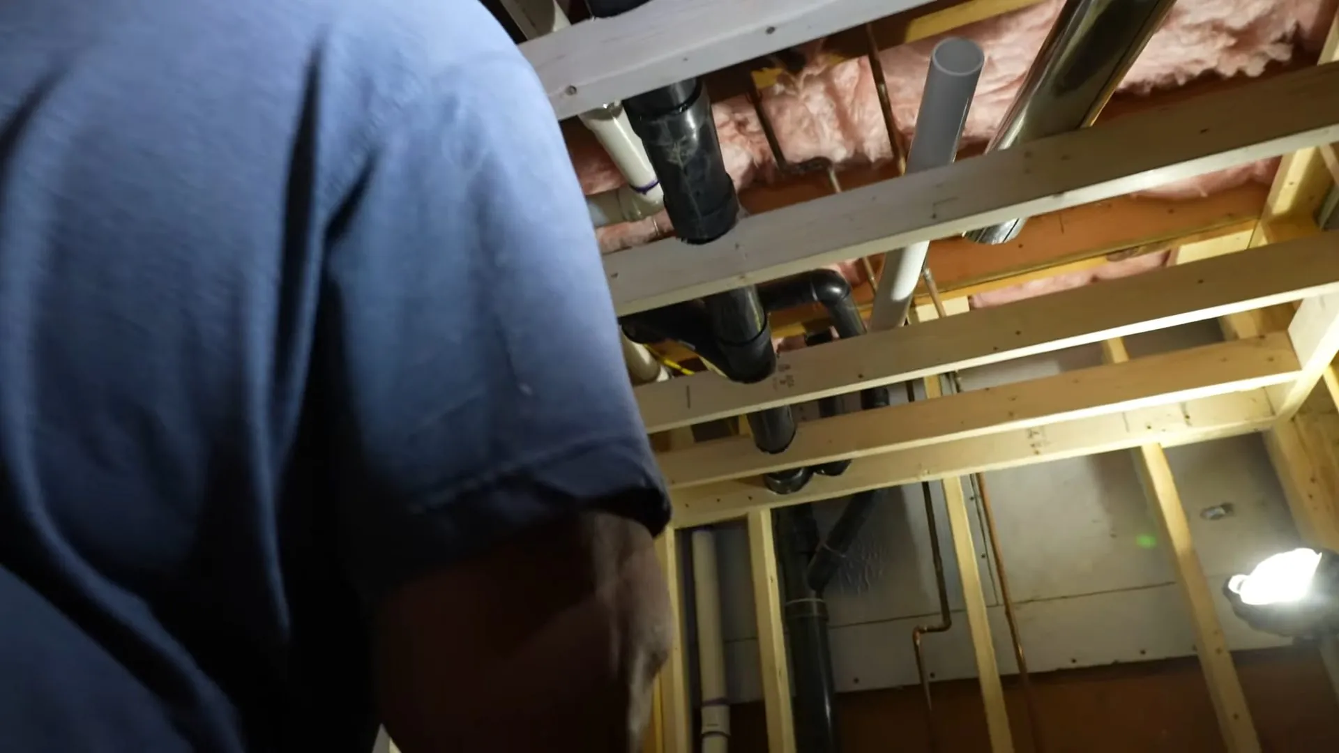 Plumber in a blue shirt working beneath basement framing with drain lines and insulation overhead