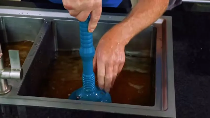 Hand working a blue plunger in a kitchen sink basin full of dirty standing water