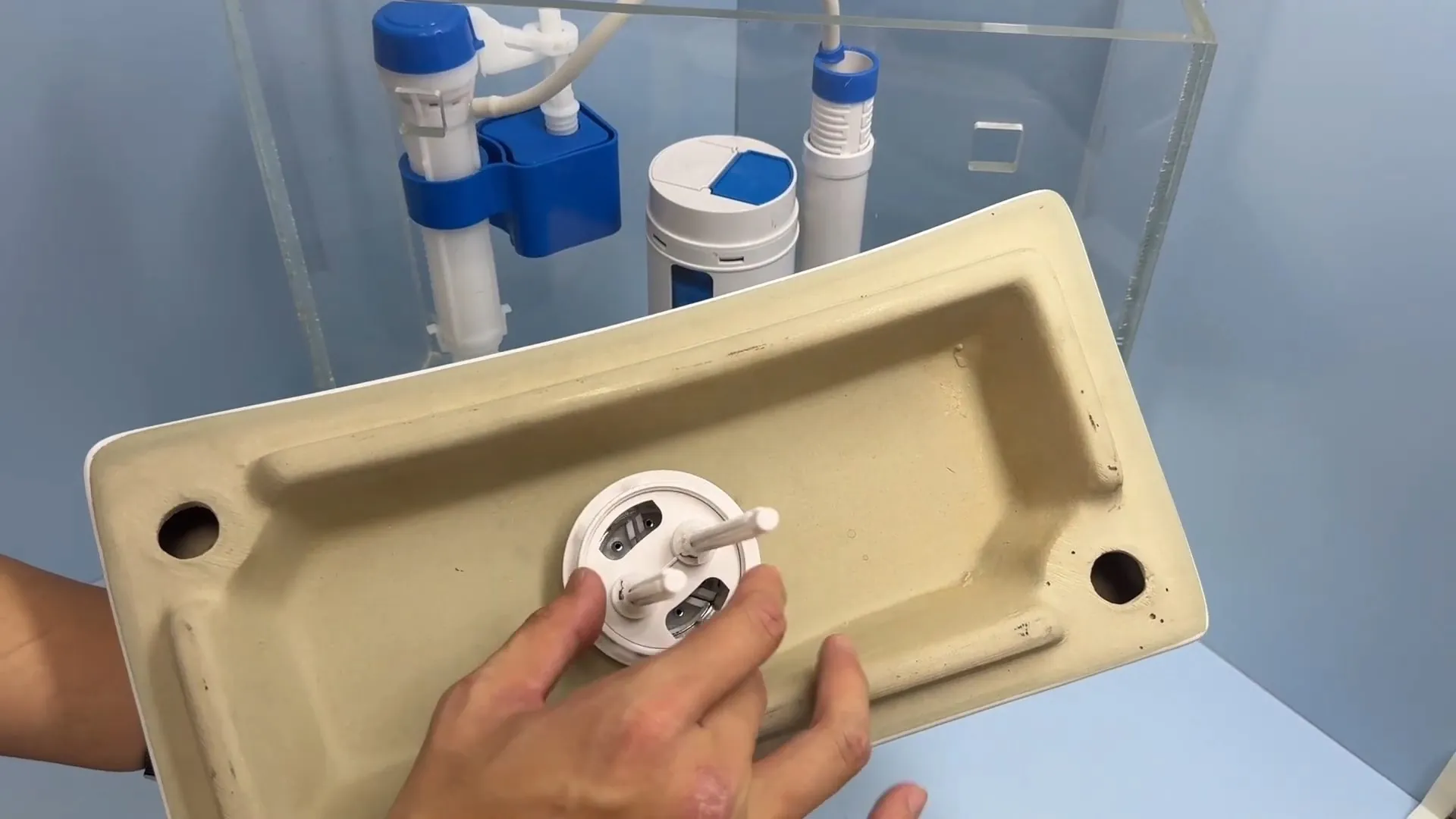 Toilet tank lid held upside down above a clear tank showing the fill valve and flapper