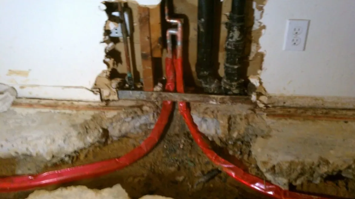Wall cut open at the baseboard with red PEX lines running through wet debris beside old pipe