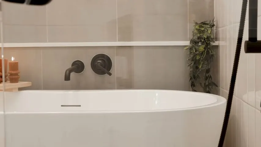 Freestanding white tub below a wall-mounted black tub filler and handle with tile behind