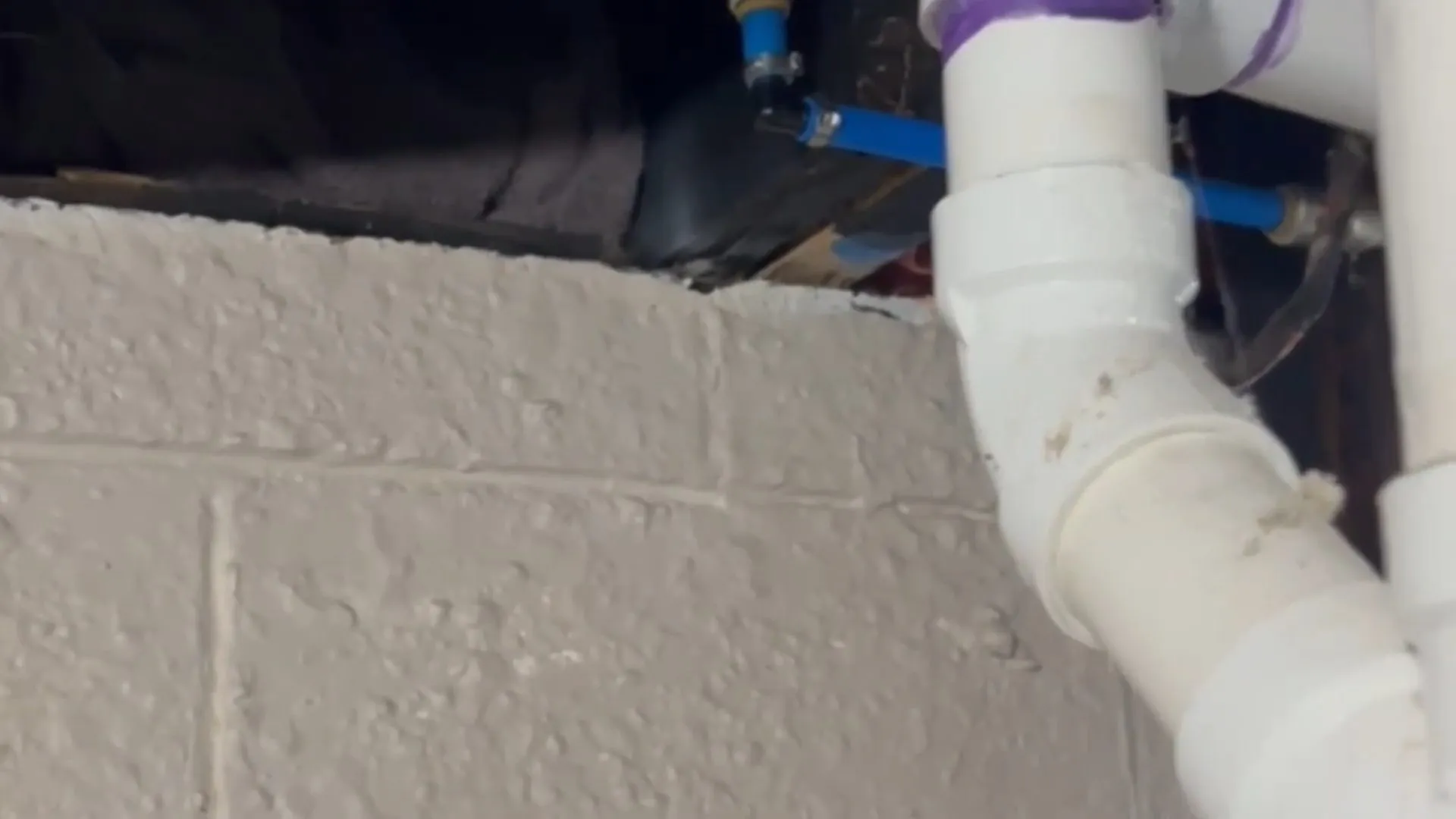 White PVC drain elbow beside a concrete block wall with blue supply tubing above