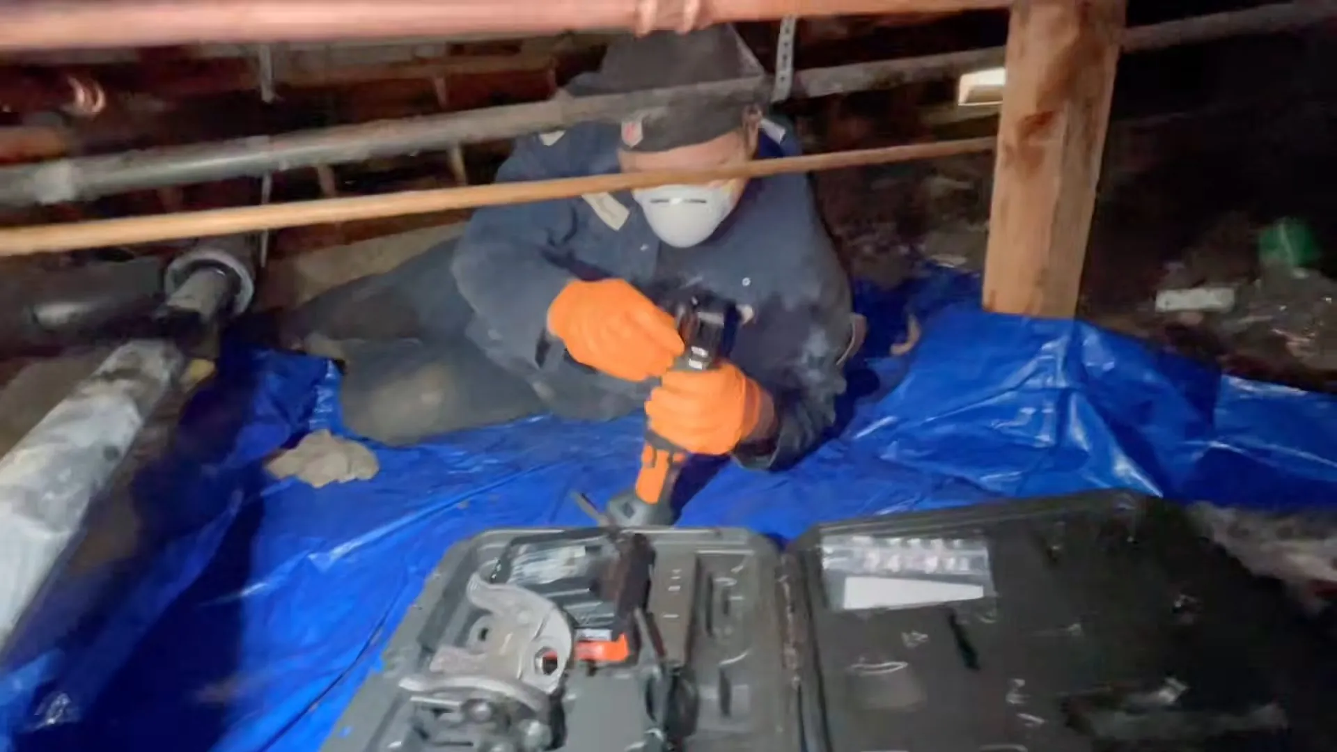 Worker kneeling on a blue tarp in a crawlspace cutting a pipe with a power tool